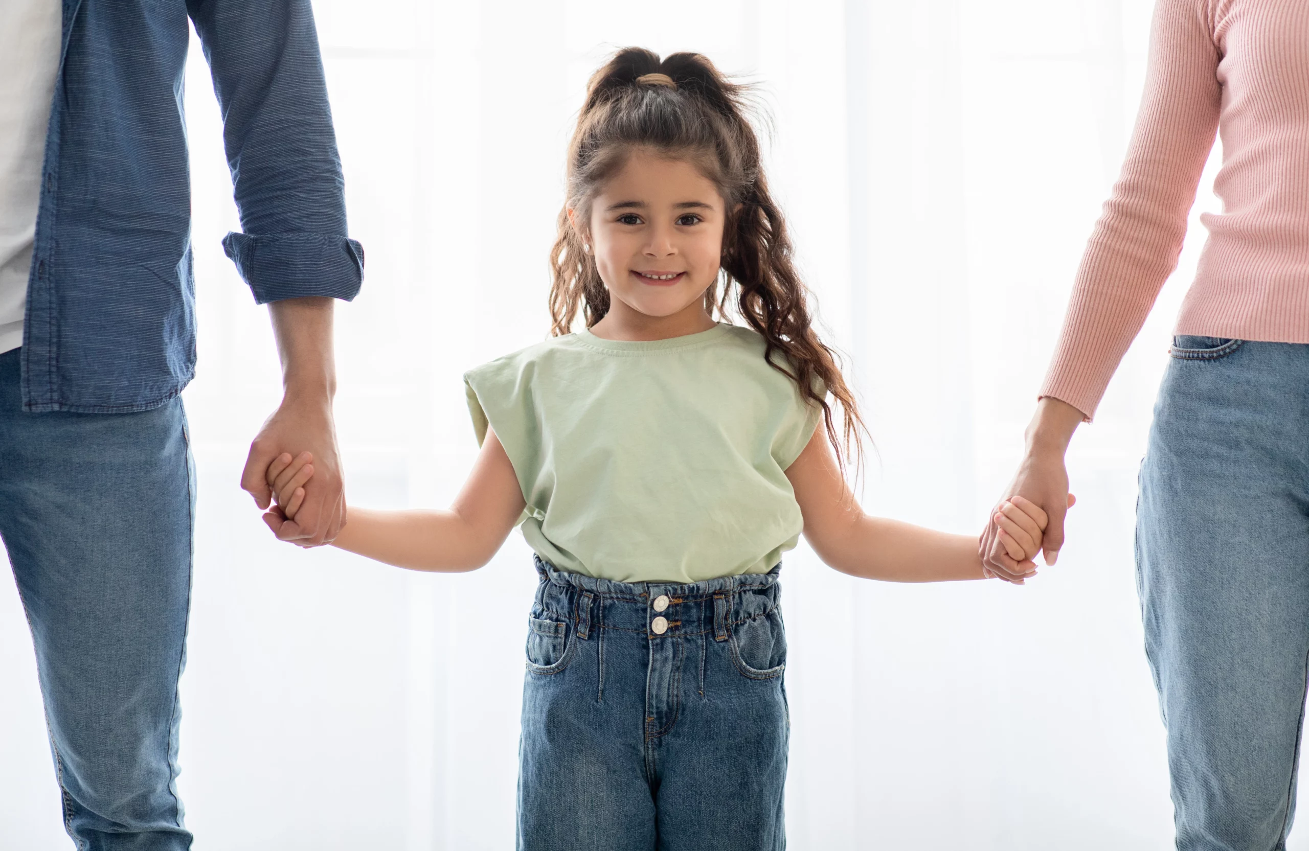 A young girl in a light green shirt and jeans smiles while holding hands with two adults, symbolizing positive co-parenting and child custody arrangements